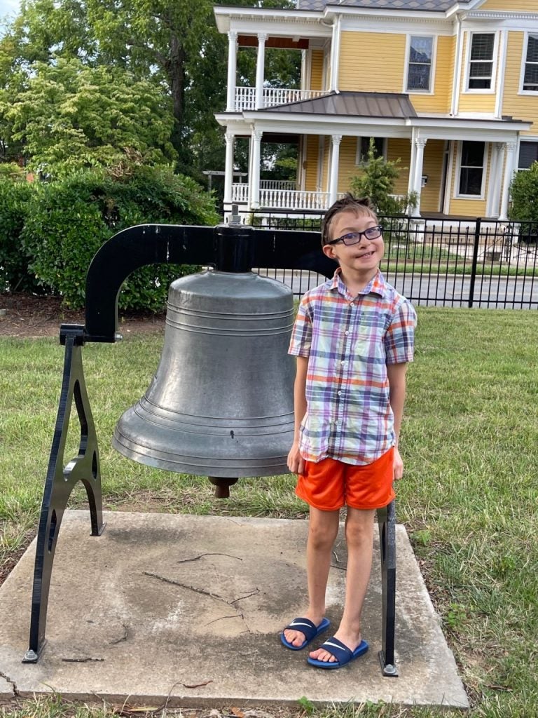 Young boy standing beside a bell. He is wearing a plaid shirt and orange shorts with navy blue Nike slides